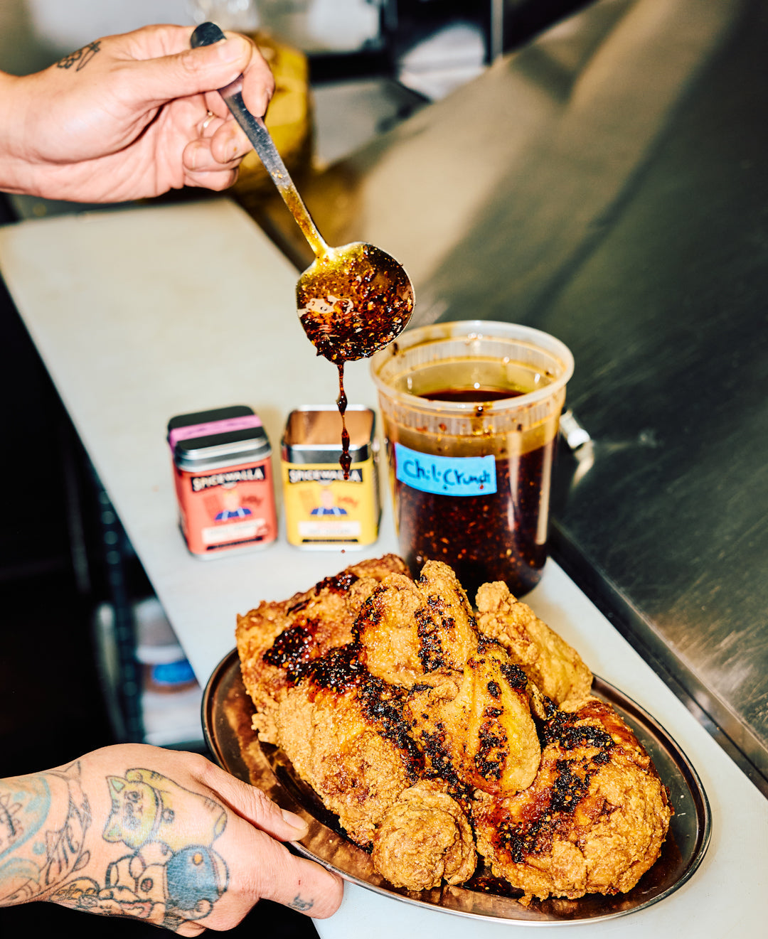 Person drizzling a sauce over fried chicken on a metal tray with a cup of coffee in the background.